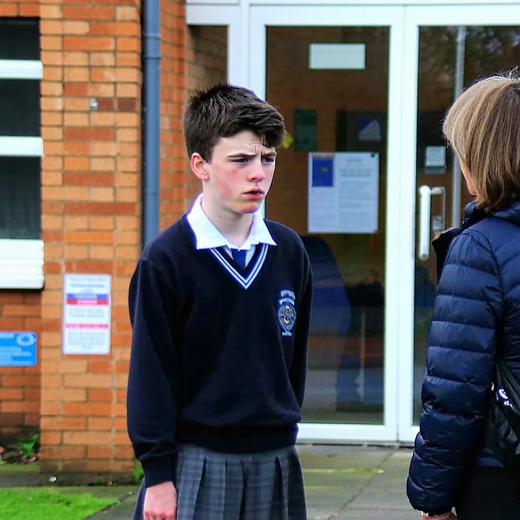 A teenager in school uniform and their parent are outside a school building from which they have just been excluded They look distressed about the decision-1